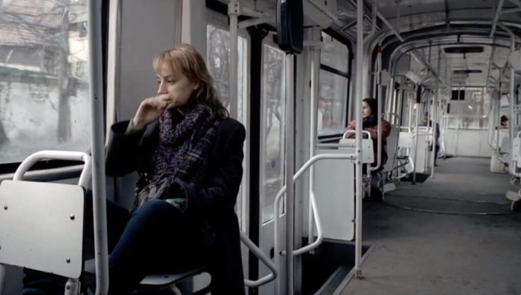 Color film still of young blonde woman in winter coat and scarf sitting on a tram.